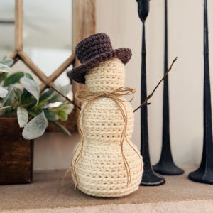May include: A handmade crochet snowman with a dark brown top hat and twig arms. The snowman is cream-colored and tied with twine. Decorative winter holiday decor.