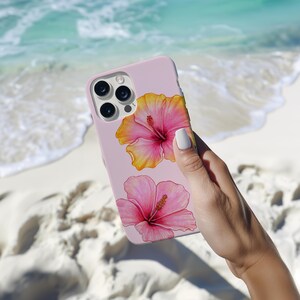 May include: A pink phone case with a floral design. The case features two hibiscus flowers, one yellow and pink and the other pink. The phone case has a camera lens cutout.