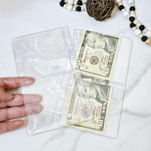 May include: Clear plastic pockets with a hole punch for a binder. Two ten dollar bills are visible inside the pockets.