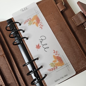 May include: A translucent plastic planner dashboard featuring the word Fall in cursive script, decorated with colorful autumn leaf illustrations and geometric lines. The dashboard is inserted into a brown textured six-ring binder.