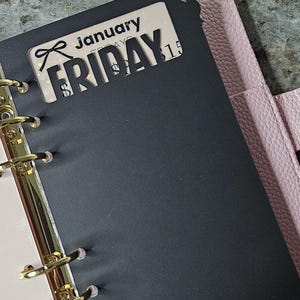 May include: A black planner dashboard with a die-cut bow at the top and the text January Friday 15. The dashboard features gold-toned metal rings and is set against a light pink textured cover. A gold safety pin with small charms is attached.
