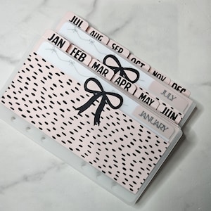 May include: A set of planner dividers featuring a pale pink background with black speckles and a prominent black bow graphic. The dividers include tabs labeled with the months of the year from January to December in black text, organized in a cascading layout.