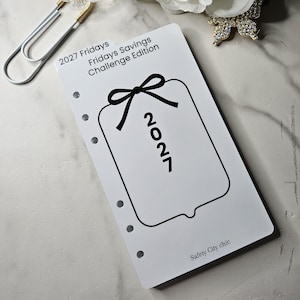 May include: A white rectangular planner insert with six punched holes along the left side. The text reads 2027 Fridays Fridays Savings Challenge Edition and Safety City chic. A black line drawing of a gift tag with a bow and the year 2027 is centered.