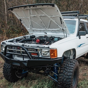 以下が含まれることがあります： フードが開いていて、エンジンが見える白いトヨタ4WDトラック。トラックには黒いバンパーと大きなオフロードタイヤが装備されています。