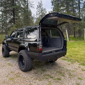 Puede incluir: Un SUV negro con suspensión elevada y neumáticos todoterreno grandes. El vehículo cuenta con un parachoques trasero negro mate, portaequipajes y un portón trasero abierto que revela un área de carga interior gris con un reposacabezas.