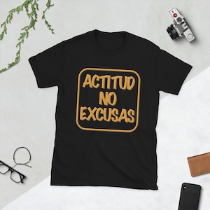 May include: Black t-shirt with the text "ACTITUD NO EXCUSAS" in a gold-colored, outlined font. The text is centered within a gold-colored, rounded rectangle. The shirt is short-sleeved and made of a soft material.