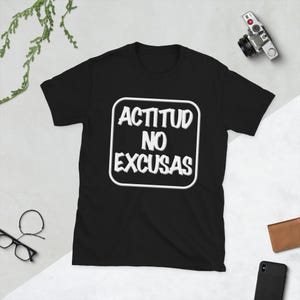 May include: Black t-shirt with the white text "ACTITUD NO EXCUSAS" inside a white outlined square. The shirt is short-sleeved and has a crew neck. A camera, glasses, a wallet, and a phone are in the background.