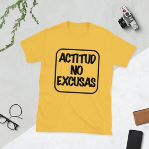 May include: A yellow t-shirt with the text "ACTITUD NO EXCUSAS" in a black, rounded rectangle. The shirt is short-sleeved and has a crew neck. The text is in a bold, sans-serif font.
