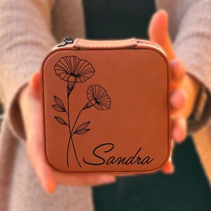 Pu&ograve; includere: Una scatola per gioielli in pelle marrone con un design floreale e il nome "Sandra" inciso sul davanti.