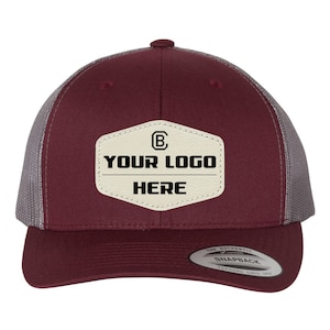 May include: A maroon and gray trucker hat with a white leather patch that says "YOUR LOGO HERE" and a small "B" inside a circle. The hat has a snap closure and the text "THE AUTHENTIC SNAPBACK YUPOONG SINCE 1974" on the back.