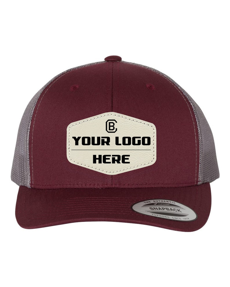 May include: A maroon and gray trucker hat with a white leather patch that says "YOUR LOGO HERE" and a small "B" inside a circle. The hat has a snap closure and the text "THE AUTHENTIC SNAPBACK YUPOONG SINCE 1974" on the back.