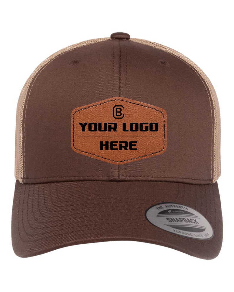 May include: A brown and tan trucker hat with a brown leather patch that says "YOUR LOGO HERE" and a letter "B" inside a circle. The hat has a mesh back and a snap closure.