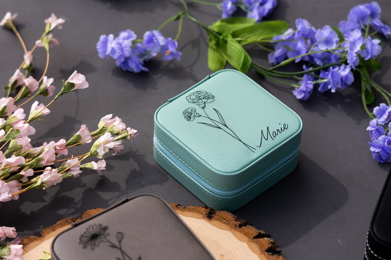 Pu&ograve; includere: Una scatola per gioielli blu turchese con chiusura a zip. La scatola ha un design floreale e il nome "Marie" inciso sulla parte superiore.
