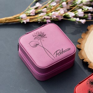 Pu&ograve; includere: Una scatola per gioielli quadrata di colore rosa con chiusura a zip. La scatola ha un design floreale e il nome "Rebecca" inciso sul davanti.