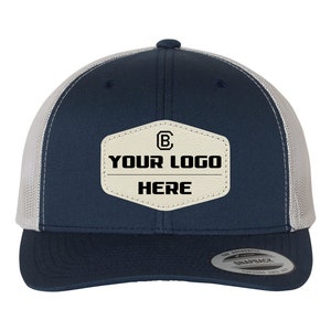 May include: A navy blue and white trucker hat with a beige leather patch that says "YOUR LOGO HERE" and a small "B" inside a circle. The hat has a snap closure and a mesh back.