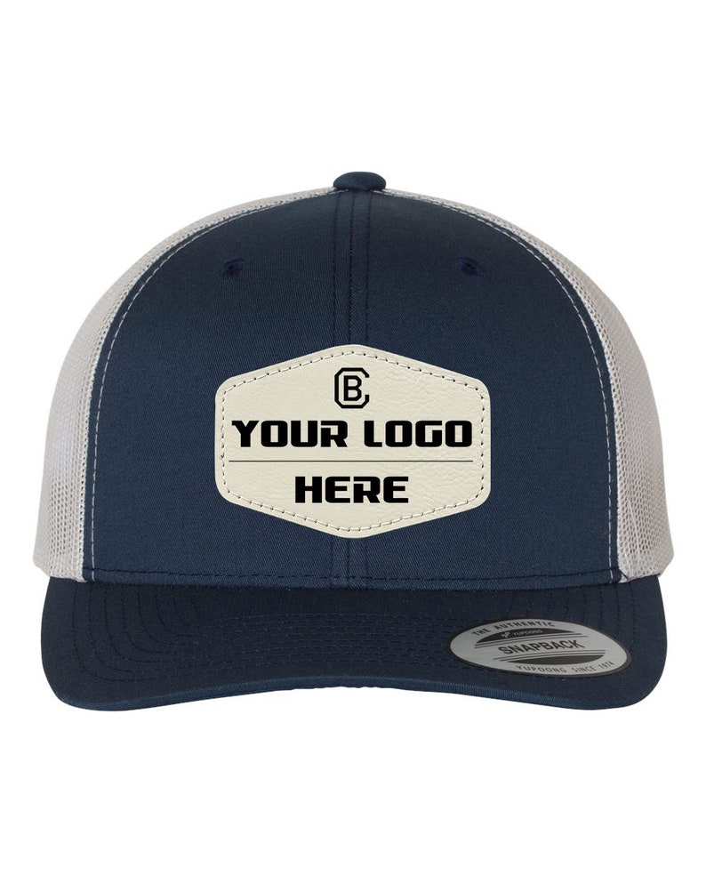 May include: A navy blue and white trucker hat with a beige leather patch that says "YOUR LOGO HERE" and a small "B" inside a circle. The hat has a snap closure and a mesh back.