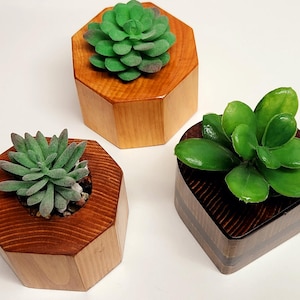 May include: Three wooden planters with faux succulents. The planters are different shapes, including a hexagon and a square. The succulents are various shades of green. A book with a blue cover is in the background.