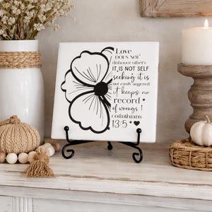 May include: A white decorative tile with a black floral design and the text "Love does not dishonor others...Corinthians 13:5" is displayed on a black metal stand. A white candle and vase are in the background.