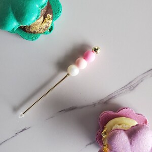 Peut inclure: Deux macarons en forme de coquillages, l'un vert et l'autre violet, avec des accents de feuilles d'or. Une brochette dorée avec des perles roses et blanches est au centre de l'image.