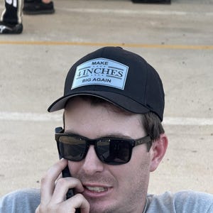 Könnte beinhalten: Schwarze Baseballkappe mit einem weißen rechteckigen Aufnäher mit der Aufschrift "MAKE 4 INCHES BIG AGAIN". Die Kappe wird von einer Person getragen, die eine Sonnenbrille trägt und telefoniert.