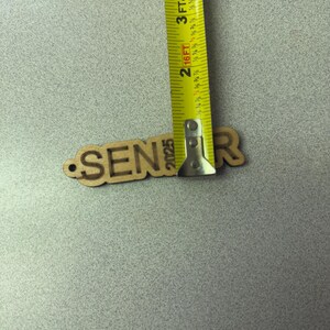 Może przedstawiać: Wycięcie z drewna słowa "Senior" z rokiem "2025" poniżej. Wycięcie jest mierzone ż&oacute;łtą miarą taśmową.