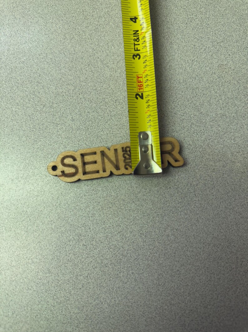 Może przedstawiać: Wycięcie z drewna słowa "Senior" z rokiem "2025" poniżej. Wycięcie jest mierzone ż&oacute;łtą miarą taśmową.