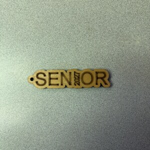 Może przedstawiać: Drewniany wycięty napis "SENIOR" z wygrawerowanym rokiem "2027" po prawej stronie.