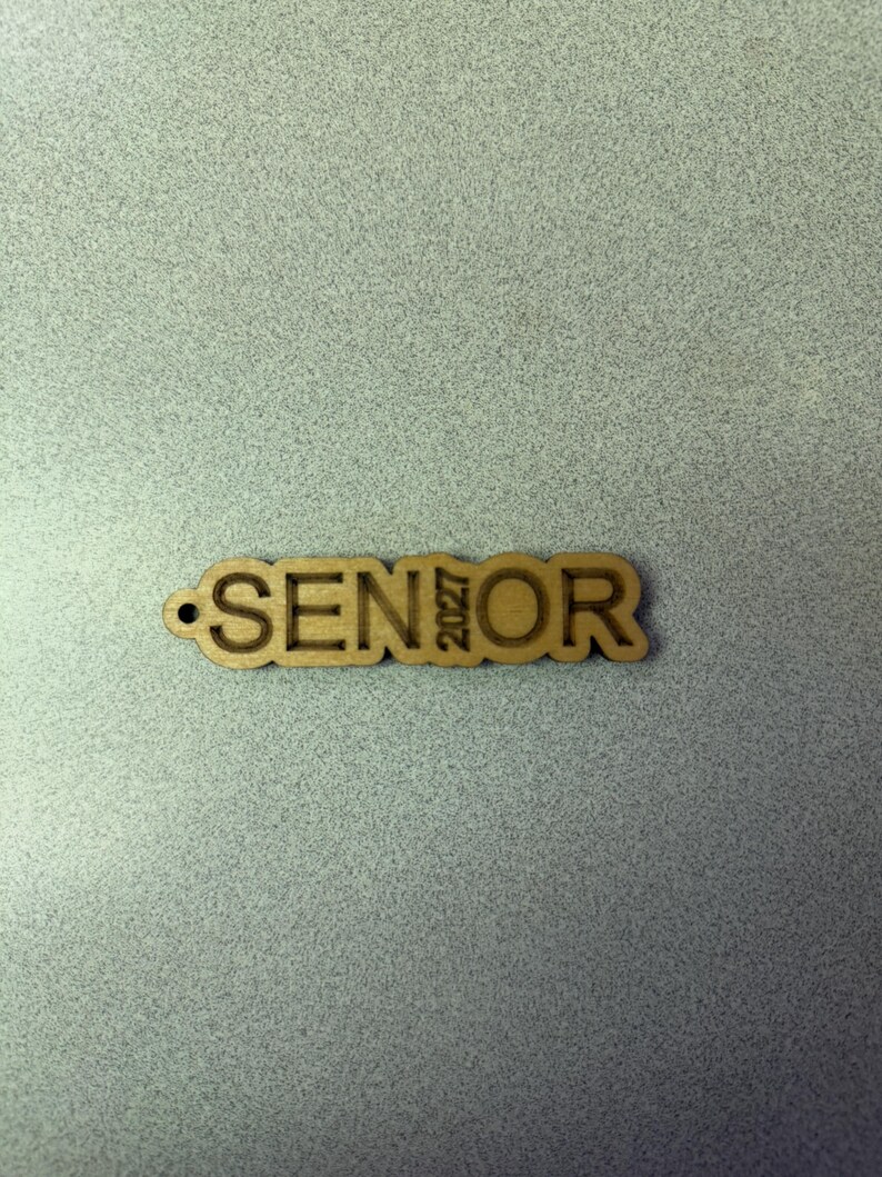 Może przedstawiać: Drewniany wycięty napis "SENIOR" z wygrawerowanym rokiem "2027" po prawej stronie.
