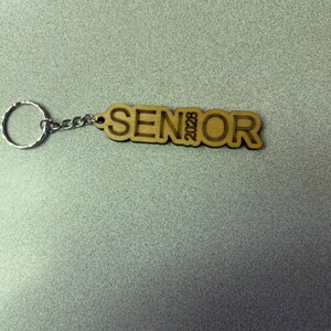 Może przedstawiać: Drewniany brelok z wygrawerowanym napisem "SENIOR 2023". Brelok jest przymocowany do srebrnego metalowego k&oacute;łka.