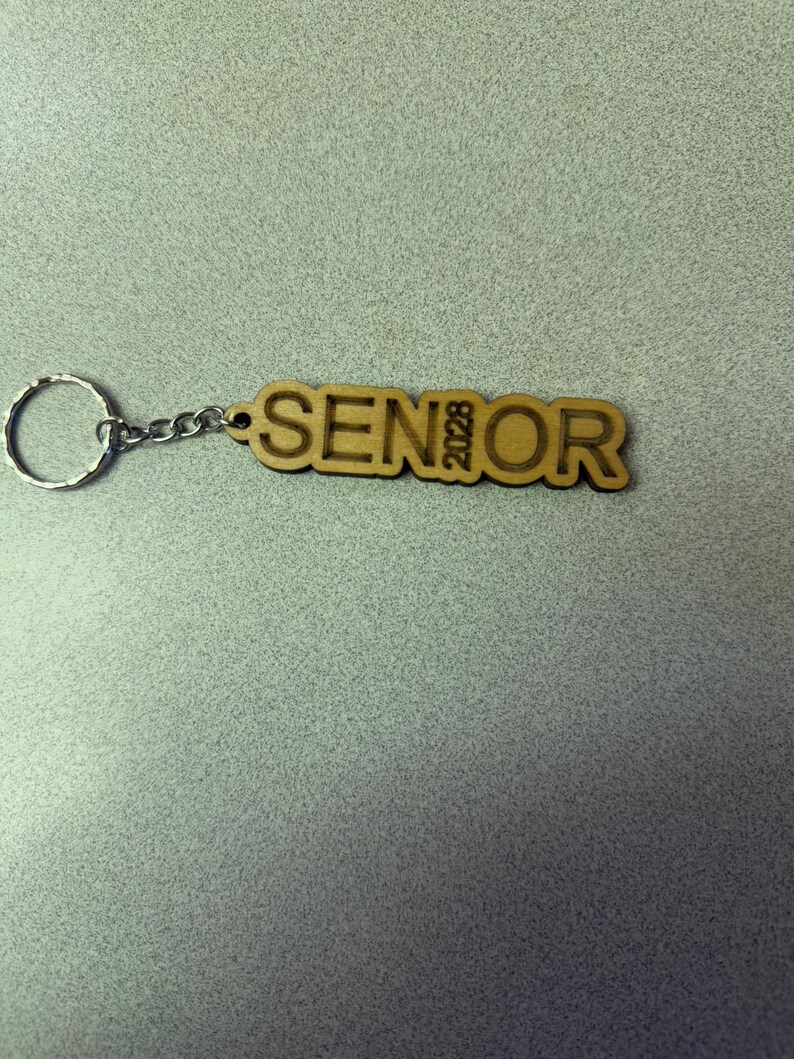 Może przedstawiać: Drewniany brelok z wygrawerowanym napisem "SENIOR 2023". Brelok jest przymocowany do srebrnego metalowego k&oacute;łka.