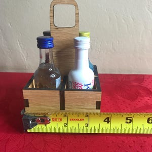 K&ouml;nnte beinhalten: Ein Holz-Flaschenhalter mit Griff. Der Halter hat vier F&auml;cher f&uuml;r Miniaturflaschen. Der Halter ist 10 cm breit. Die Flaschen sind aus klarem Glas mit blauen, wei&szlig;en und gelben Kappen.