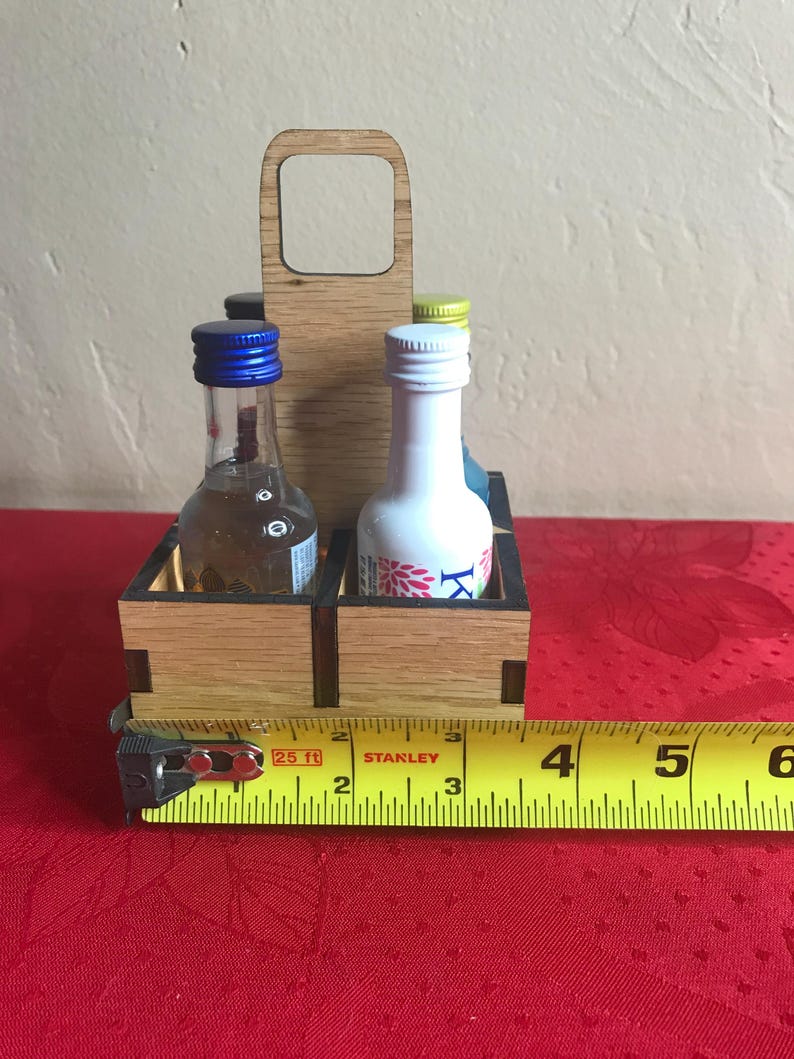 K&ouml;nnte beinhalten: Ein Holz-Flaschenhalter mit Griff. Der Halter hat vier F&auml;cher f&uuml;r Miniaturflaschen. Der Halter ist 10 cm breit. Die Flaschen sind aus klarem Glas mit blauen, wei&szlig;en und gelben Kappen.