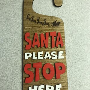 Könnte beinhalten: Ein hölzerner Türhänger mit einem Weihnachtsmann-Motiv. Der Hänger ist braun mit roten und weißen Buchstaben, auf denen "Santa Please Stop Here" steht.