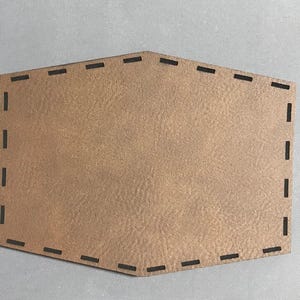 May include: A brown leather patch with a decorative stitched border. The patch is shaped like a hexagon with rounded corners.