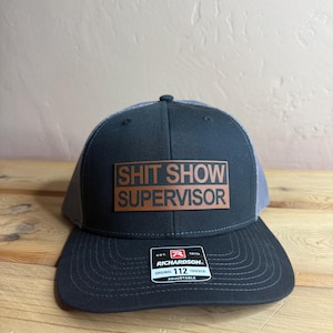 May include: Black and grey trucker hat with a brown leather patch that says "Shit Show Supervisor". The hat is adjustable and has a Richardson 112 label.