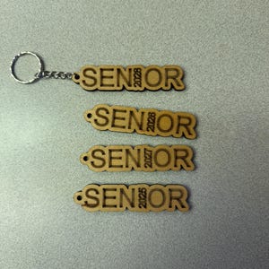 Może przedstawiać: Cztery drewniane breloki z wygrawerowanym napisem "SENIOR" i rokiem 2028, 2026, 2027 i 2025.