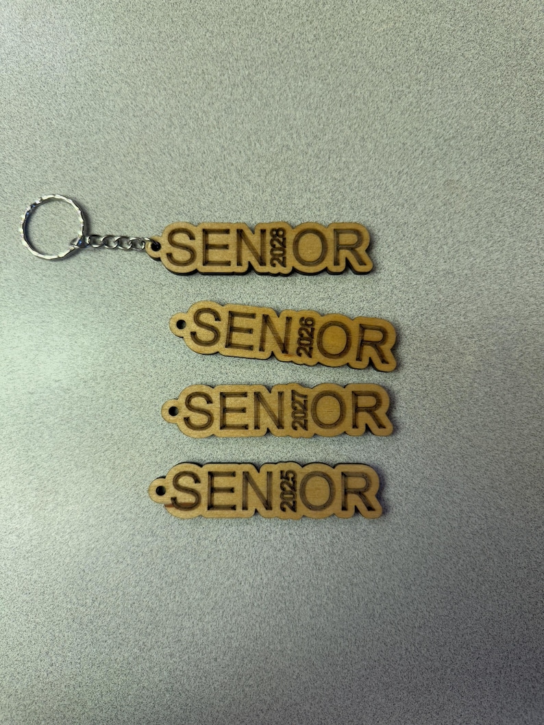 Może przedstawiać: Cztery drewniane breloki z wygrawerowanym napisem "SENIOR" i rokiem 2028, 2026, 2027 i 2025.