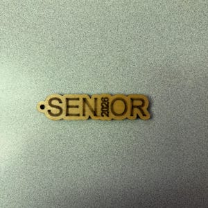 Może przedstawiać: Drewniany brelok do kluczy z wygrawerowanym napisem "SENIOR" i rokiem "2023".