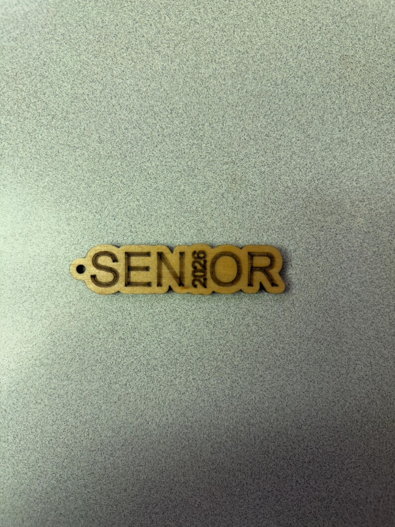 Może przedstawiać: Drewniany brelok do kluczy z wygrawerowanym napisem "SENIOR" i rokiem "2023".