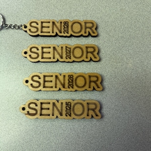 Może przedstawiać: Cztery drewniane breloki z napisem "SENIOR" i rokiem 2025, 2026, 2027 i 2028.