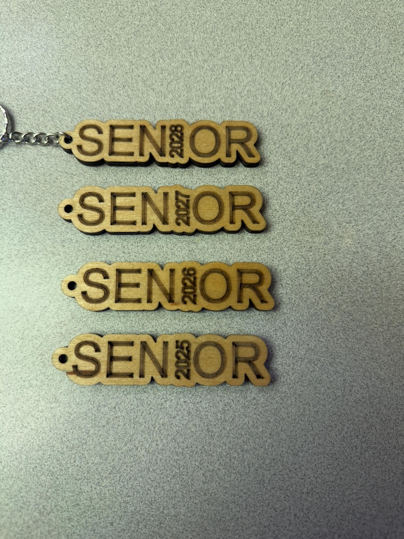 Może przedstawiać: Cztery drewniane breloki z napisem "SENIOR" i rokiem 2025, 2026, 2027 i 2028.