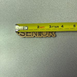 Może przedstawiać: Drewniany znak z napisem "Senior" i rokiem "2023". Znak jest mierzony ż&oacute;łtą miarą.