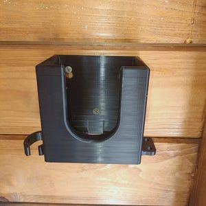 Puede incluir: Un soporte de pared negro de forma rectangular impreso en 3D, con un recorte frontal en forma de U, ganchos laterales y bridas de montaje. El soporte est&aacute; fijado a una superficie de madera con dos tornillos met&aacute;licos visibles en la cavidad.