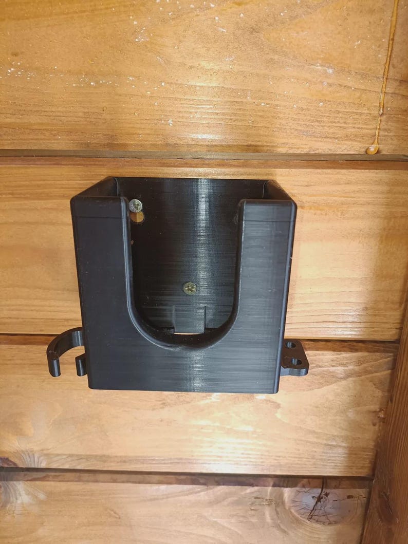 Puede incluir: Un soporte de pared negro de forma rectangular impreso en 3D, con un recorte frontal en forma de U, ganchos laterales y bridas de montaje. El soporte est&aacute; fijado a una superficie de madera con dos tornillos met&aacute;licos visibles en la cavidad.