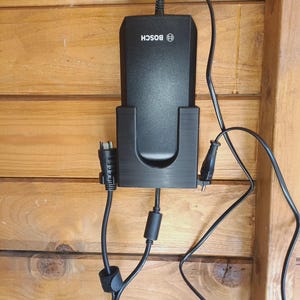 Puede incluir: Un cargador de bater&iacute;a Bosch negro montado en un soporte de pl&aacute;stico negro. El cargador cuenta con un cable de alimentaci&oacute;n superior y dos cables negros adicionales que se extienden por los lados y la parte inferior, con un n&uacute;cleo de ferrita.