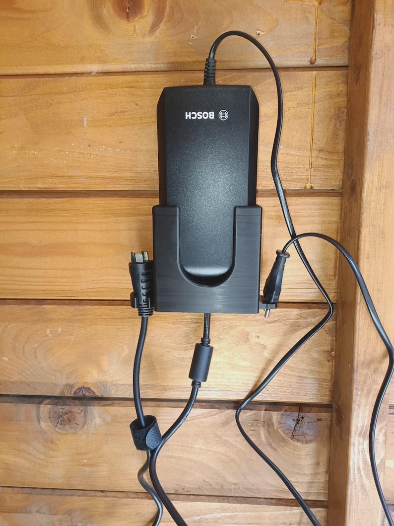 Puede incluir: Un cargador de bater&iacute;a Bosch negro montado en un soporte de pl&aacute;stico negro. El cargador cuenta con un cable de alimentaci&oacute;n superior y dos cables negros adicionales que se extienden por los lados y la parte inferior, con un n&uacute;cleo de ferrita.