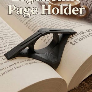 Könnte beinhalten: Ein schwarzer ergonomischer Seitenhalter liegt auf einem offenen Buch. Der Seitenhalter mit strukturierter Oberfläche hat einen Ring für den Finger. Das Buch ist auf einer Seite mit spanischem Text geöffnet.