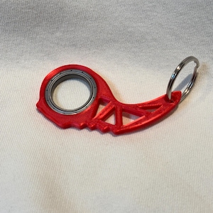 May include: A red 3D-printed keychain with a bearing in the center and a key ring attached. The keychain has a unique, geometric design with triangular cutouts. The object is set against a white background.