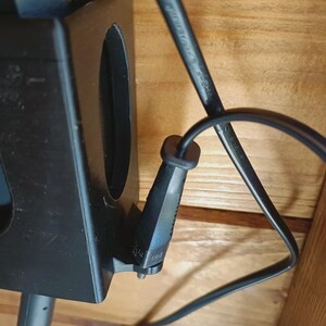 Puede incluir: Un soporte de pl&aacute;stico negro impreso en 3D con una ranura vertical y un clip lateral para cables. Un cable de alimentaci&oacute;n negro est&aacute; insertado en el clip, mientras que un segundo cable negro sale de la parte inferior del soporte en madera.