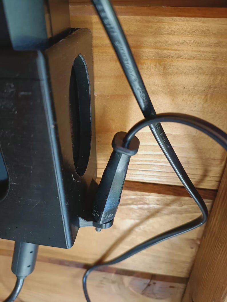 Puede incluir: Un soporte de pl&aacute;stico negro impreso en 3D con una ranura vertical y un clip lateral para cables. Un cable de alimentaci&oacute;n negro est&aacute; insertado en el clip, mientras que un segundo cable negro sale de la parte inferior del soporte en madera.